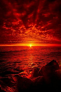 the sun is setting over the ocean with rocks in the foreground and red clouds above it