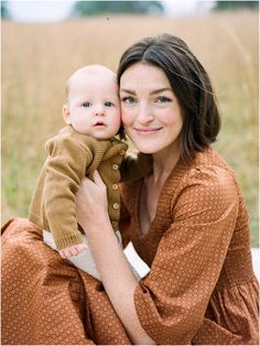 a woman holding a baby in her arms while sitting on the ground with tall grass behind her