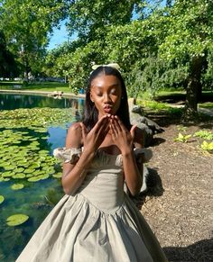 a girl in a dress is standing by the water with her hands clasped to her chest