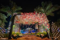 the entrance to an outdoor event decorated with lights and flowers, surrounded by palm trees