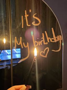 a person holding a lit candle in front of a mirror that says it's my birthday