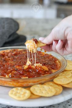 a person dipping cheese into a bowl of chili dip surrounded by crackers