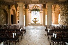 rows of chairs with white bows are lined up in front of an ornate fountain and columns