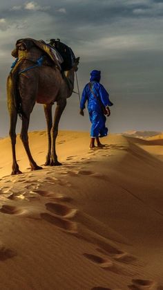 a man in blue is walking with a camel on the sand dunes, while another person walks behind him