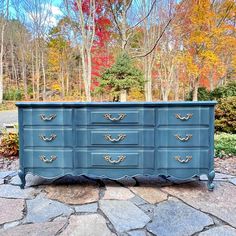 a blue dresser sitting on top of a stone floor in front of trees with leaves