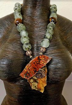 Homage to Mother Earth! Two glossed slabs of jasper (Morgan Poppy and Ocean) host an ammonite fossil as well as burnt orange crystals in this OOAK unisex statement pendant!  Flanking the pendant are two ornate copper beads.  The necklace portion consists of African recycled glass beads which are a frosted white with brown spots. Two oversized deep chocolate brown beads with ornate silver tone star bead caps serve as accents.  The back is finished with tiger's eye.  Closes with gold tone hardware Luxury Statement Necklace With Large Stone, Luxury Large Beaded Necklaces As Statement Piece, Handmade Artisan Ocean Jasper Necklace, Unique Handmade Jasper Necklace, Handmade Jasper Artisan Beaded Necklaces, Handmade Jasper Beaded Necklaces Artisan Style, Handmade Jasper Beaded Necklaces, Handmade Unique Jasper Beaded Necklaces, Unique Ocean Jasper One-of-a-kind Necklace