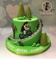 a green birthday cake with a woman riding a bike on the road and trees around it