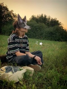 a person sitting in the grass with a cat mask on their head and a bag