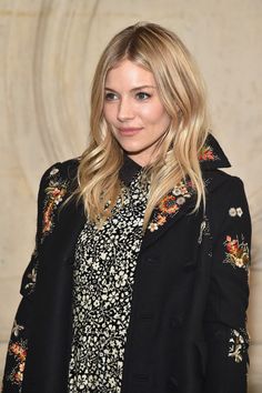 a woman standing in front of a wall wearing a black jacket and floral dress shirt