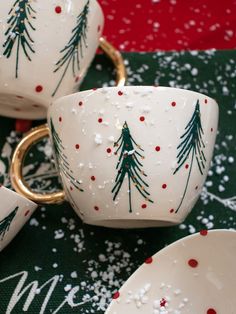 two white cups with christmas trees painted on them and gold rimmed mugs sitting next to each other