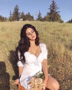 a woman sitting in the grass with a basket full of flowers on her lap and looking at the camera