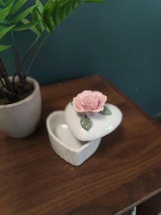 a small pink rose sitting on top of a white heart shaped dish next to a potted plant