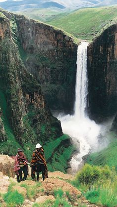 two people sitting on the edge of a cliff looking at a large waterfall in the distance