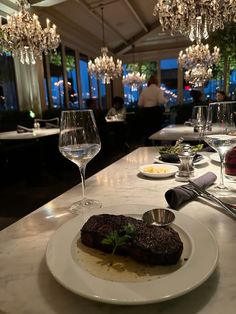 a white plate topped with meat next to glasses of wine and plates of food on a table