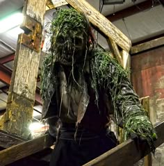 a scarecrow dressed in green and black standing next to a wooden cross with moss growing on it