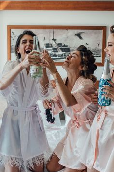 three bridesmaids in robes toasting and laughing while sitting on a white bed