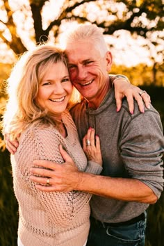 an older man and woman hugging each other in front of the camera with trees behind them