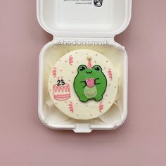 a cake shaped like a frog sitting in a white container with pink frosting on it