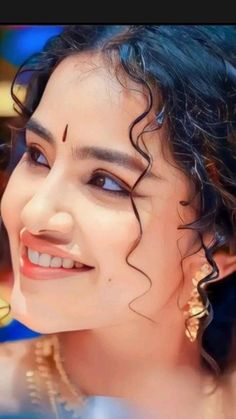 a woman with curly hair and blue eyes smiles at the camera while wearing gold jewelry