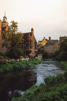 a river running through a lush green forest filled with trees and tall brick buildings on either side of it