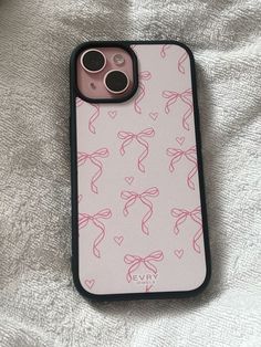 an iphone case with pink bows on it sitting on a white bed sheet, next to a black cell phone