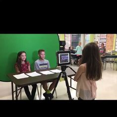 two children are sitting at a table in front of a green screen while someone is recording them