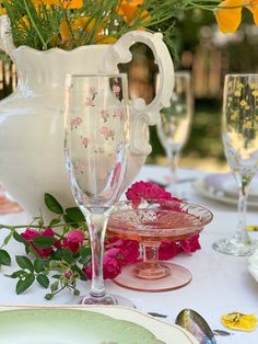 the table is set with glasses, plates and flowers in vases on top of it