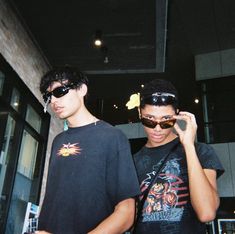 two young men standing next to each other in front of a building and one is wearing sunglasses