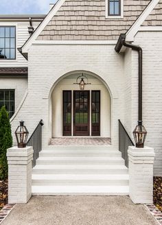 the front entrance to a white brick house with two lanterns on each door and steps leading up to it