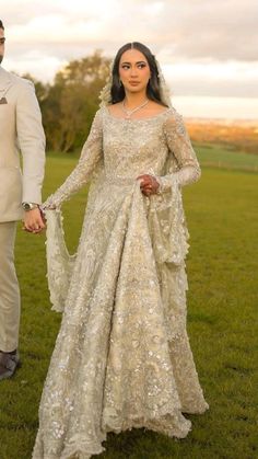 a man and woman dressed in wedding attire holding hands while standing on green grass with trees in the background