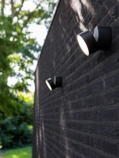 two lights on the side of a brick wall next to a green lawn and trees