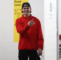 a young man in a red jacket and black cap holding a water bottle while standing next to a white wall