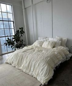 an unmade bed with white ruffled sheets and pillows in front of a large window