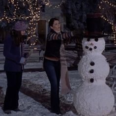 two women standing next to a snowman in the middle of a yard with christmas lights