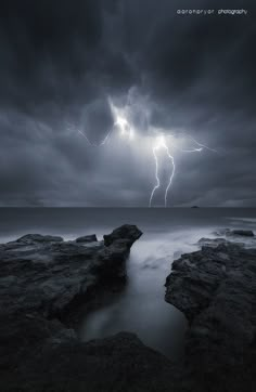 a storm is coming over the ocean with rocks in front of it and lightning above