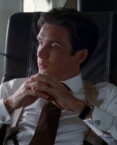 a man wearing a white shirt and brown tie sitting in a chair with his hands on his chin