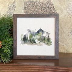 a watercolor painting of mountains and trees on a shelf next to pine cones with evergreen needles in the foreground