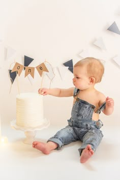 a baby in overalls sitting next to a cake with one on it and some bunting flags