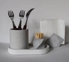 three forks and two napkins in a cup on a white surface with other utensils