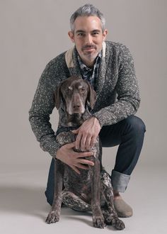 a man kneeling down next to a dog on a gray background with his legs crossed