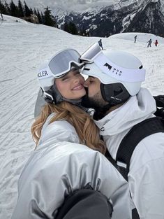 a man and woman kissing in the snow with skis on their heads, wearing goggles