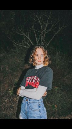 a woman standing in front of a tree at night with her arms crossed and looking off to the side