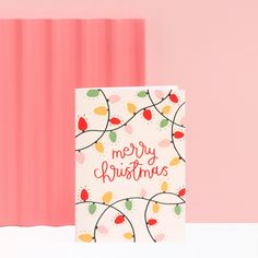 a christmas card sitting on top of a table next to a pink wall with red, green and yellow lights