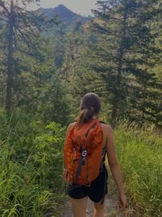 a woman walking up a trail in the woods with an orange backpack on her back