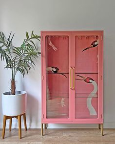 a pink cabinet with birds on it next to a potted plant