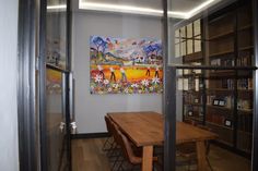 a wooden table sitting in front of a book shelf next to a painting on the wall