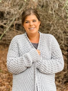 a woman standing in front of bushes wearing a gray cardigan and smiling at the camera