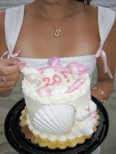 a woman is holding a cake decorated with seashells and pink bows on the beach