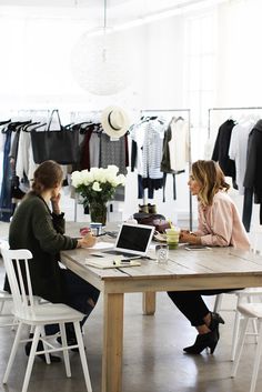 two women sitting at a table working on their laptops in a clothing store,