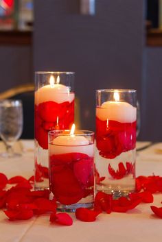 three candles with rose petals in them on a table next to two glasses filled with water